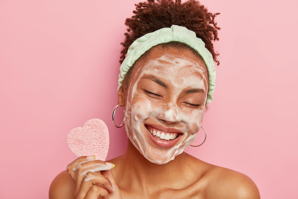 African American woman cleanses face with foam, refreshes skin, has well cared complexion, holds heart shaped sponge to represent the ways to show your skin some love